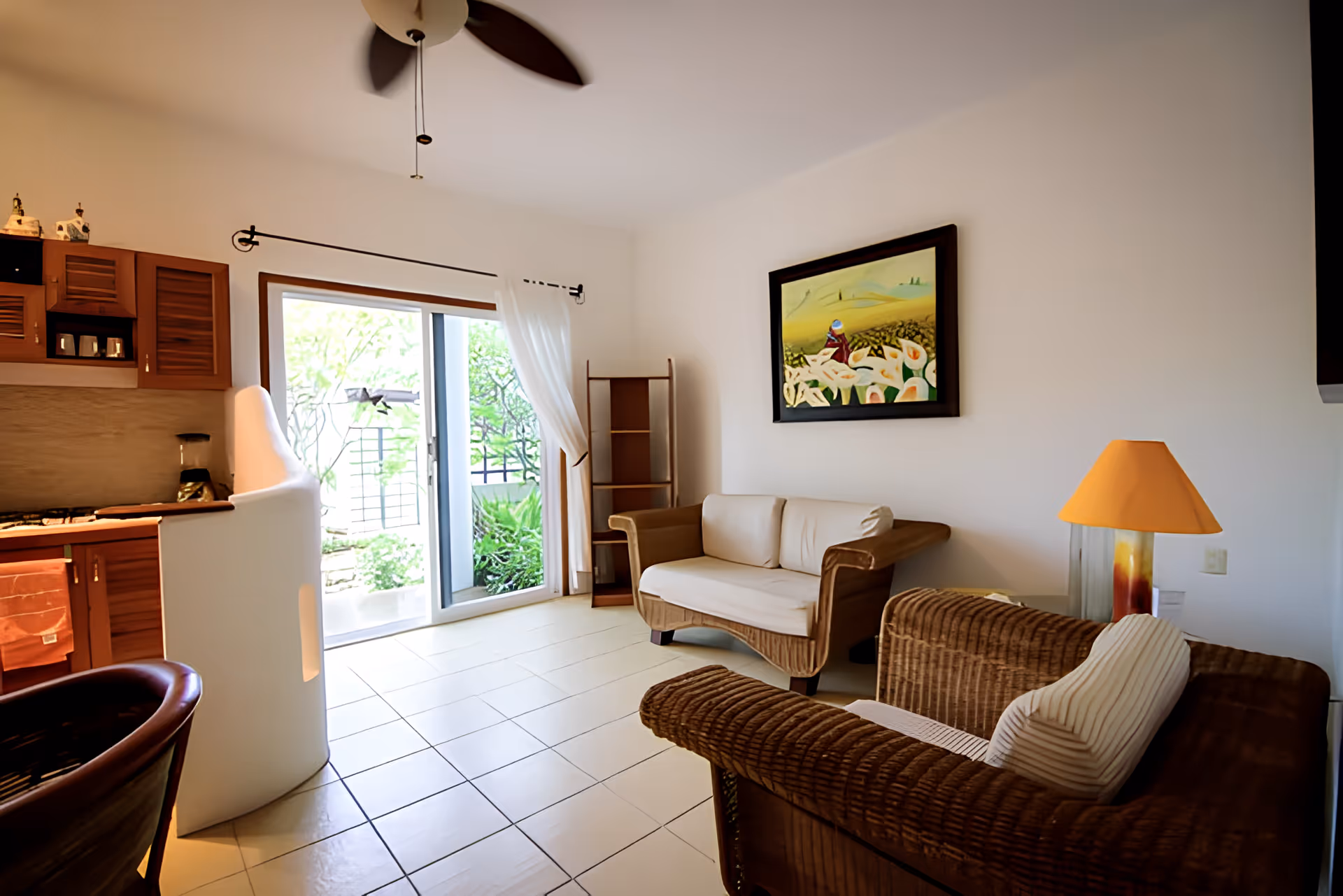 A bright living room with wicker furniture including a sofa and armchair, a small kitchen area with wooden cabinets, a ceiling fan, a painting on the wall, and a sliding glass door leading to a garden outside.