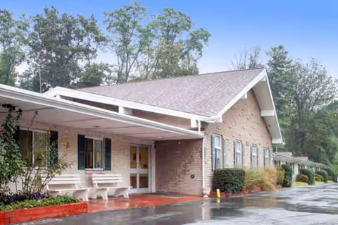 Single-story brick healthcare building with a covered entrance, benches, and a wet driveway surrounded by trees.