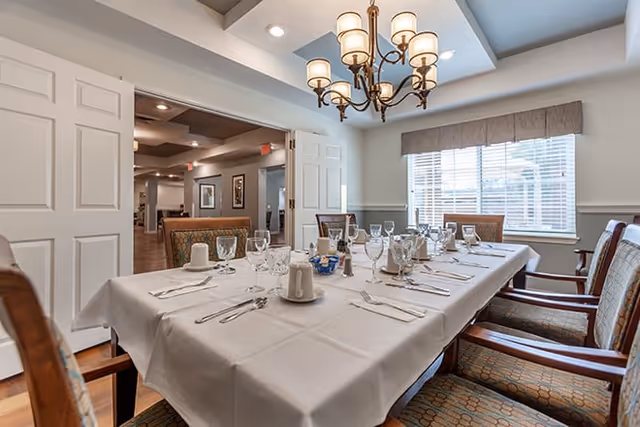 A dining room with a long table covered with a white tablecloth set for a meal, including plates, glasses, napkins, and silverware. The room has upholstered chairs around the table, a window with blinds and a valance, and a chandelier hanging from the ceiling. The walls are light-colored with wainscoting, and there is an open doorway leading to another room.