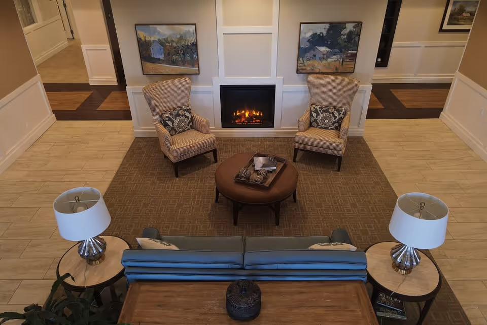 A cozy senior living facility common area with two patterned armchairs facing a lit fireplace, a round ottoman in the center, a blue sofa with two side tables each holding a lamp, and framed landscape paintings on the wall.