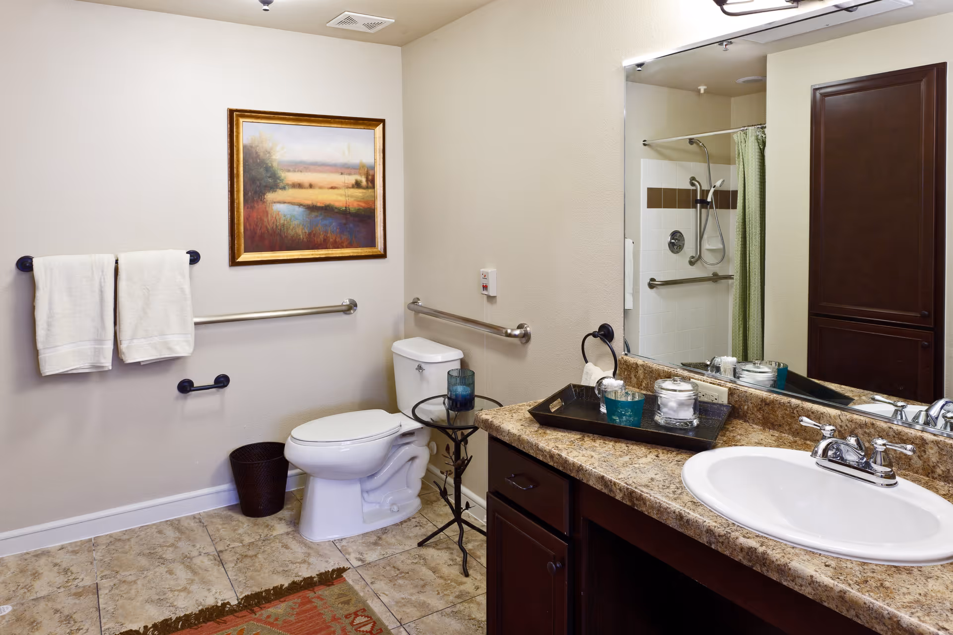 A clean and accessible bathroom featuring a toilet with grab bars on the walls, a small side table with a blue glass, a countertop with a sink, and a large mirror above it. There are two white towels hanging on a towel rack, a framed landscape painting on the wall, and a shower with a green curtain visible in the mirror's reflection.