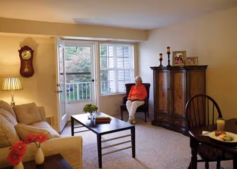 An elderly woman sits in a living room with a sofa, table, and wooden cabinet.