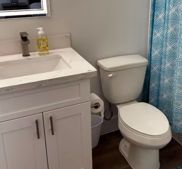 A bathroom with a white vanity cabinet topped with a marble countertop and an integrated sink. A modern faucet and a bottle of hand soap are on the countertop. Next to the vanity is a white toilet, and to the right, a blue shower curtain with a white geometric pattern is visible.