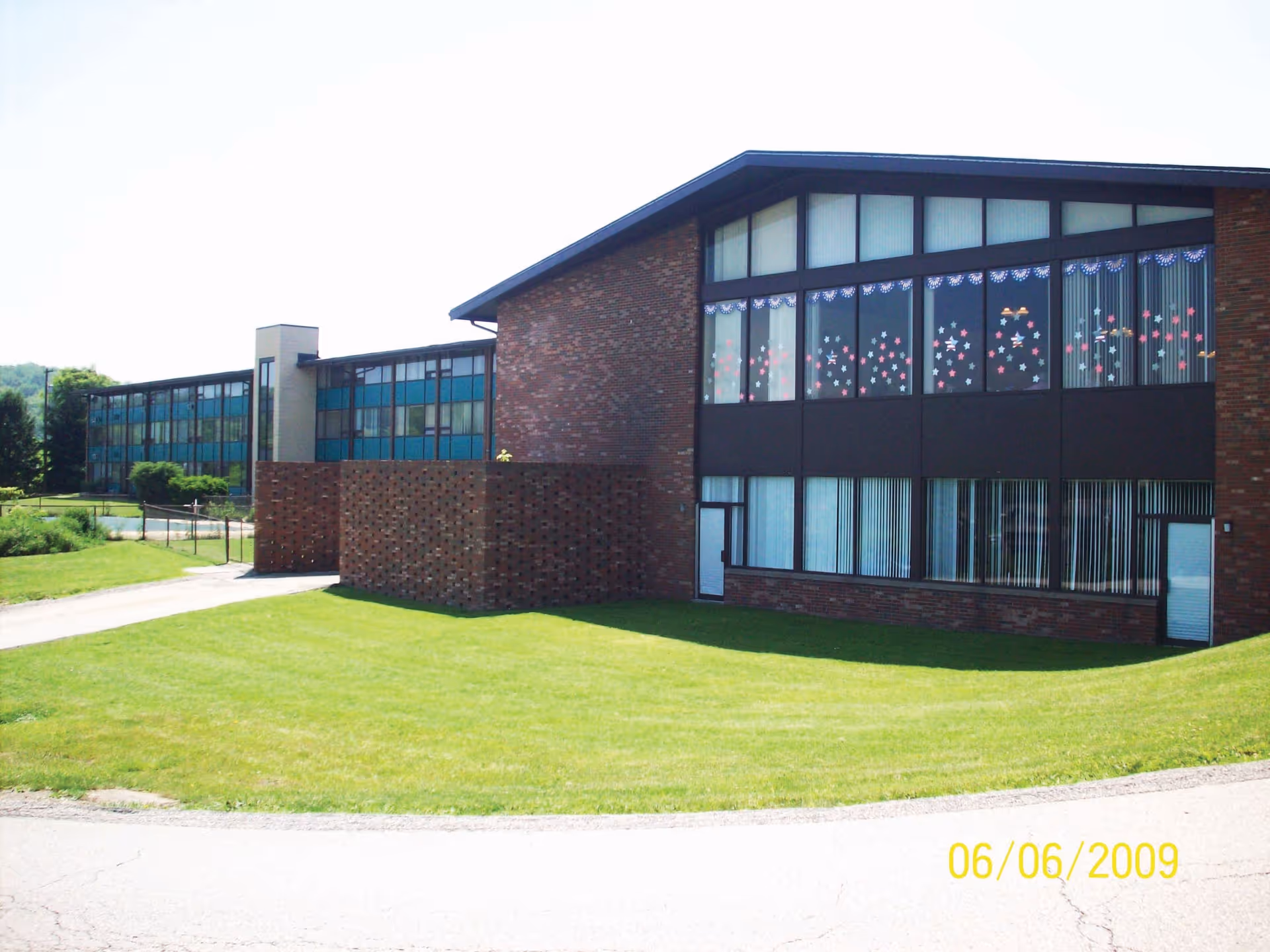 Exterior view of a two-story brick building with large windows decorated with star-shaped stickers and patriotic bunting. The building is surrounded by a well-maintained green lawn and a paved driveway. The sky is clear and bright.