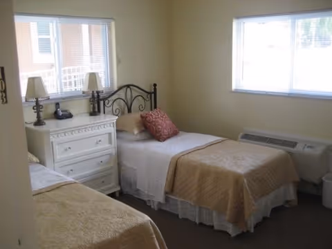 A bedroom with two single beds, each with beige bedspreads and white sheets. Between the beds is a white dresser with two lamps and a telephone on top. The room has two windows with blinds, beige walls, and a heating/cooling unit beneath one window.