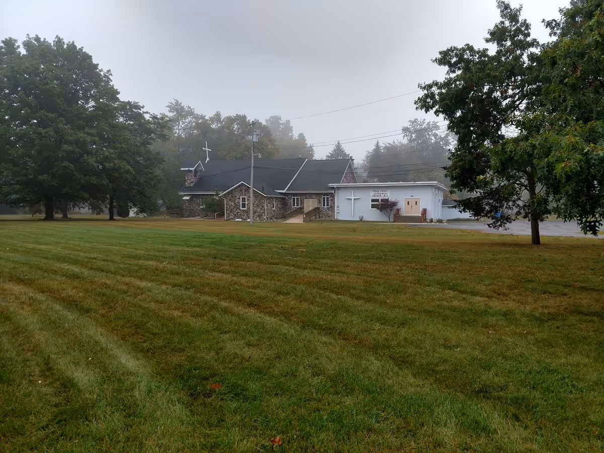 A large grassy lawn with a few trees in the foreground and a stone and white building with crosses on it in the background, under a foggy sky.