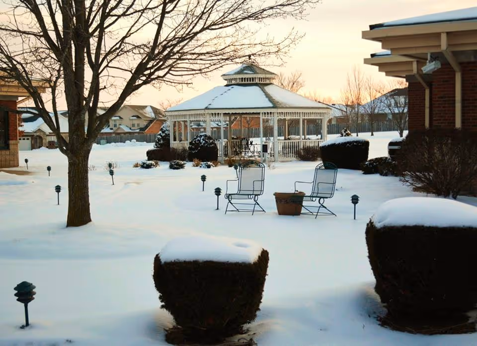 Snow-covered outdoor area at Baptist Homes of Ozark featuring a white gazebo, leafless trees, trimmed bushes topped with snow, two metal chairs, and a small table, all under a soft evening sky.