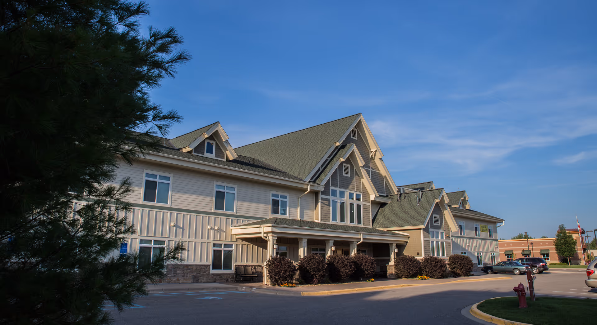 Sunlit senior living facility with peaked roofs, shrubs, and a parking lot.