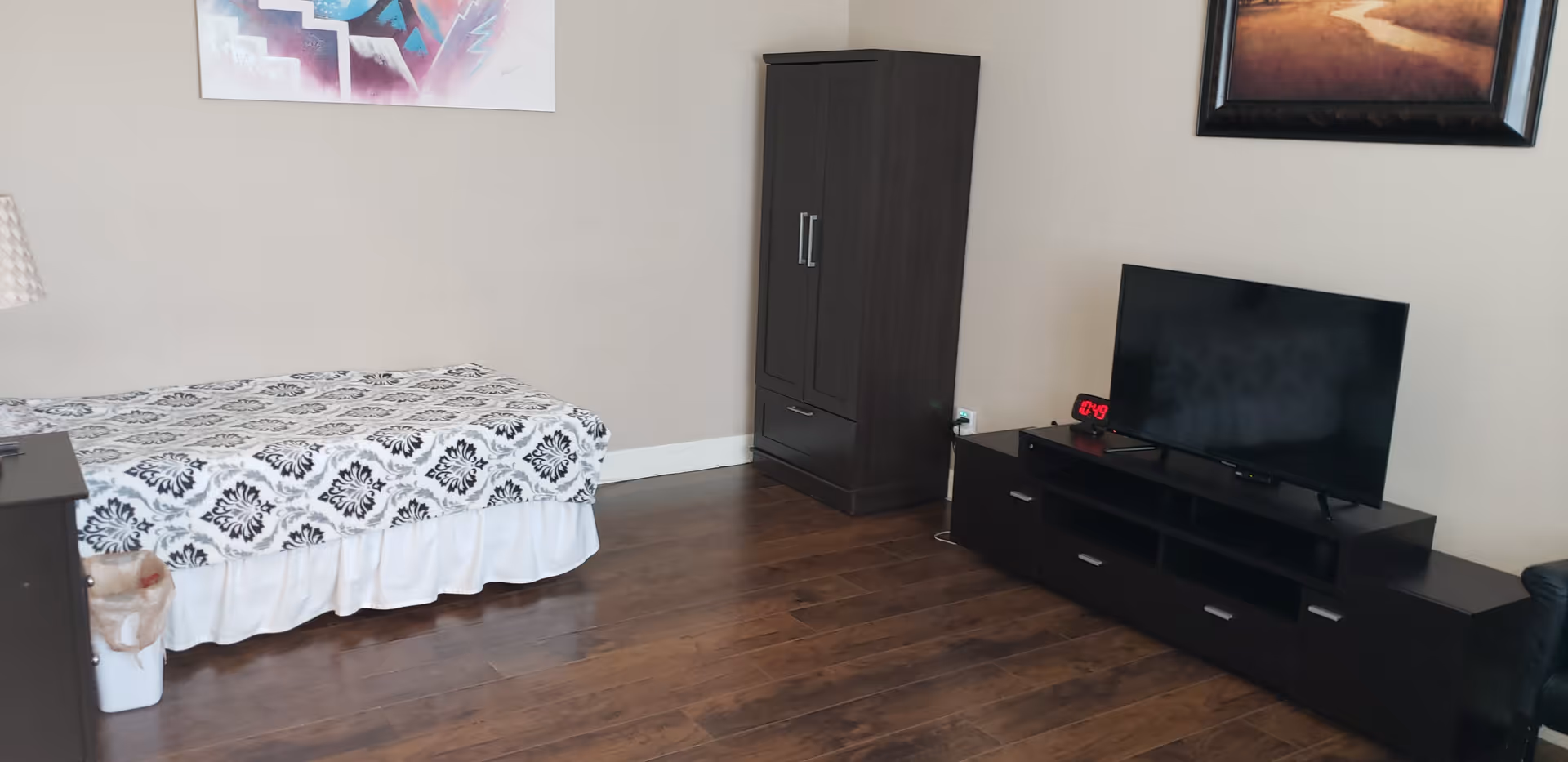 A bedroom with a single bed covered in a white and black patterned bedspread, a dark wooden wardrobe, a matching TV stand with a flat-screen TV, a digital clock, and a framed picture on the wall. The room has wooden flooring and neutral-colored walls.