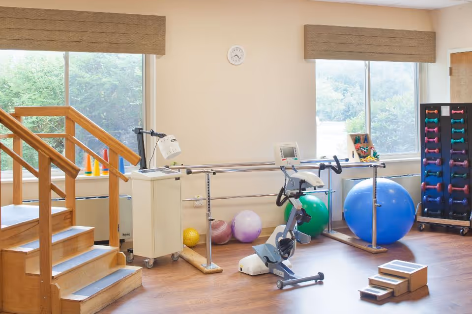 A rehabilitation room with wooden stairs for physical therapy, an exercise bike, parallel bars, various exercise balls, colorful dumbbells on a rack, and a small set of wooden steps. The room has large windows with beige blinds letting in natural light.