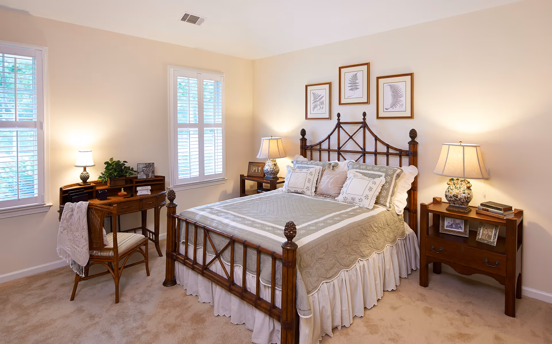 Well-lit traditional bedroom with a wooden bed, bedside tables and lamps, a small desk and two shuttered windows.