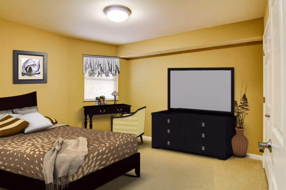 A bedroom with beige walls and carpet, featuring a bed with patterned brown and beige bedding and pillows. There is a small desk with books and a lamp under a window with a decorative valance. A chair is placed next to the desk. Across from the bed is a black dresser with a large mirror or TV on top, and a decorative vase with tall, thin branches beside it. A framed flower picture hangs on the wall above the bed.