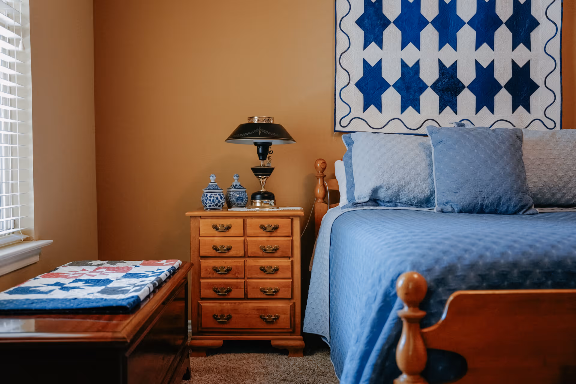 A cozy bedroom featuring a wooden bed with blue bedding and pillows, a wooden nightstand with a black lamp and decorative blue and white jars, a wooden chest with a folded quilt on top, and a wall hanging quilt with blue star patterns.