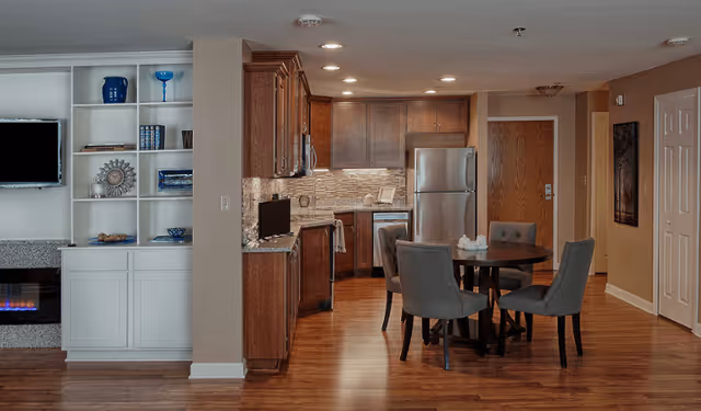 Interior view of a senior living facility unit featuring a kitchen with wooden cabinets, stainless steel refrigerator, and a small round dining table with four gray upholstered chairs. To the left, there is a built-in white shelving unit with decorative items and a wall-mounted flat-screen TV above a modern electric fireplace. The floor is wooden, and the walls are painted beige.