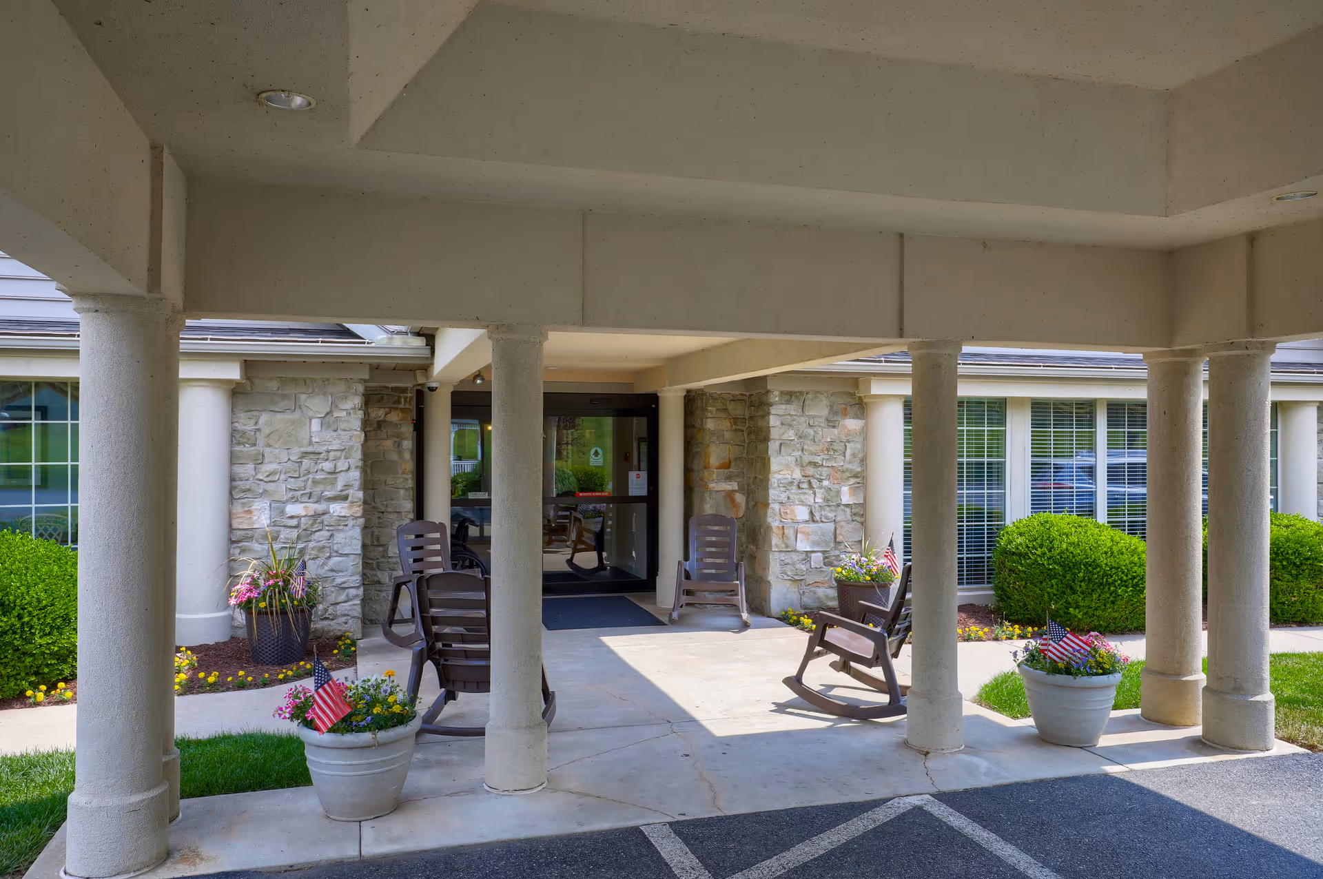 Covered entrance area of a senior living facility with stone pillars and walls, several brown rocking chairs, potted plants with flowers and small American flags, and glass doors leading inside.