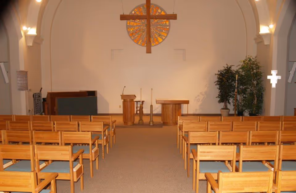 Interior view of a chapel with wooden pews arranged in rows facing a wooden altar and podium. A large wooden cross hangs on the wall above a circular stained glass window. There are potted plants on the right side and soft lighting on the walls.