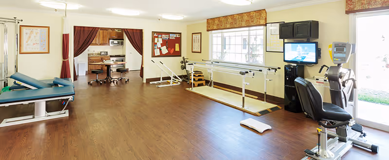 A spacious rehabilitation room with wooden flooring featuring physical therapy equipment including a treatment table, parallel bars for walking exercises, a stationary exercise bike with a screen, and a set of stairs for mobility training. The room has large windows with floral valances allowing natural light, a bulletin board on the wall, and an open doorway with curtains leading to another room with additional equipment.