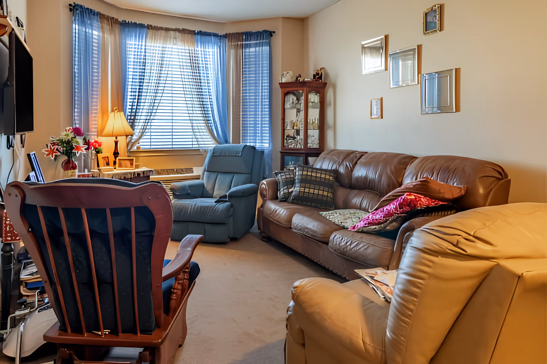 A cozy living room with a brown leather sofa adorned with colorful pillows, a blue recliner chair near a window with blue and beige curtains, a wooden rocking chair with dark cushions, a beige armchair, a small side table with a lamp and flowers, and a glass-front wooden cabinet in the corner. Several framed pictures and mirrors hang on the beige walls.