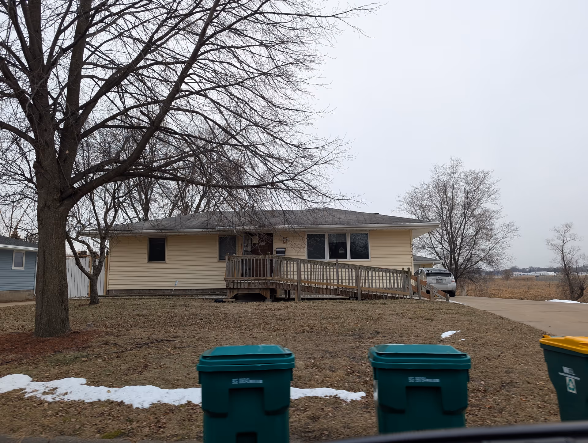 Single-story beige house with a wooden wheelchair ramp leading to the front door. Leafless trees surround the house and patches of snow are visible on the ground. Three green and yellow trash bins are in the foreground near the street.