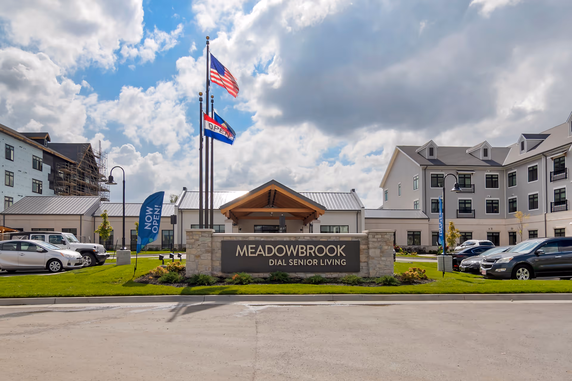 Front exterior view of Meadowbrook Dial Senior Living facility with a large sign in front, three flagpoles with American and open flags, parked cars, and a partly cloudy sky.