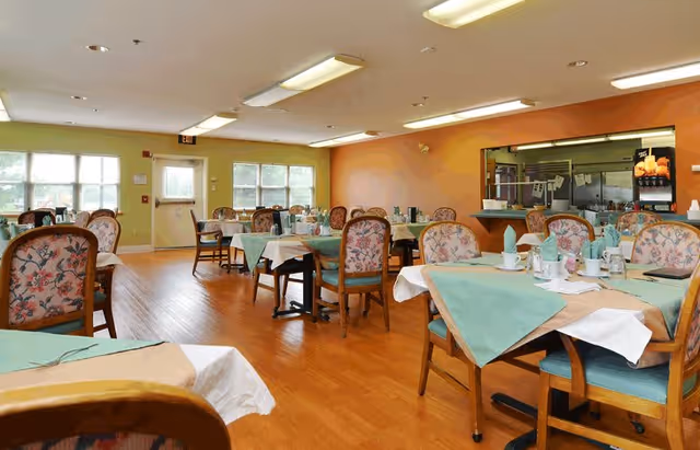 A dining room in a senior living facility with several tables covered with white and green tablecloths. Each table is set with cups, napkins, and utensils. The chairs have floral upholstery. The room has wooden flooring, large windows letting in natural light, and a serving window to the kitchen area.