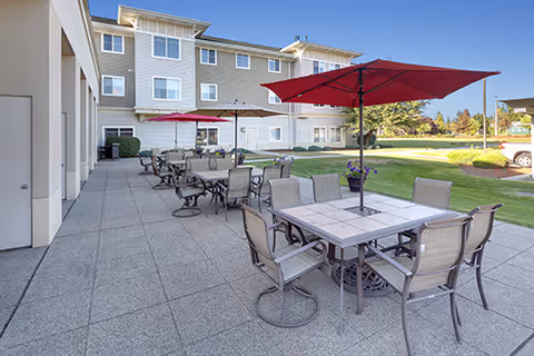 Outdoor patio with multiple dining tables and red umbrellas beside a multi-story senior living building and lawn.