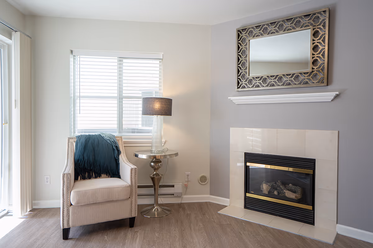 A bright living room corner with an upholstered armchair draped with a throw, a glass lamp on a round side table, and a tiled fireplace topped by a decorative mirror.