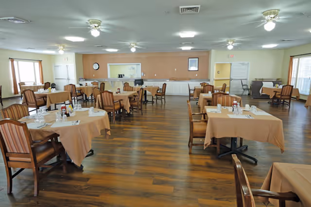 A spacious dining room with multiple tables covered in beige tablecloths, each set with napkins, glasses, and condiments. The room has wooden flooring, striped cushioned chairs, ceiling fans with lights, and large windows allowing natural light to enter.