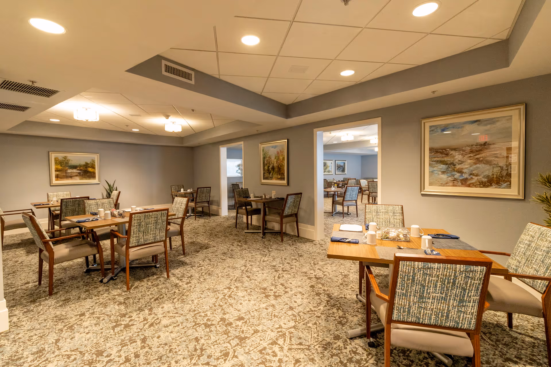 A spacious dining room with multiple wooden tables and cushioned chairs arranged neatly. The room has soft lighting from ceiling fixtures, light gray walls adorned with framed landscape paintings, and a patterned carpeted floor. Some tables are set with placemats, napkins, cups, and small decorative items.