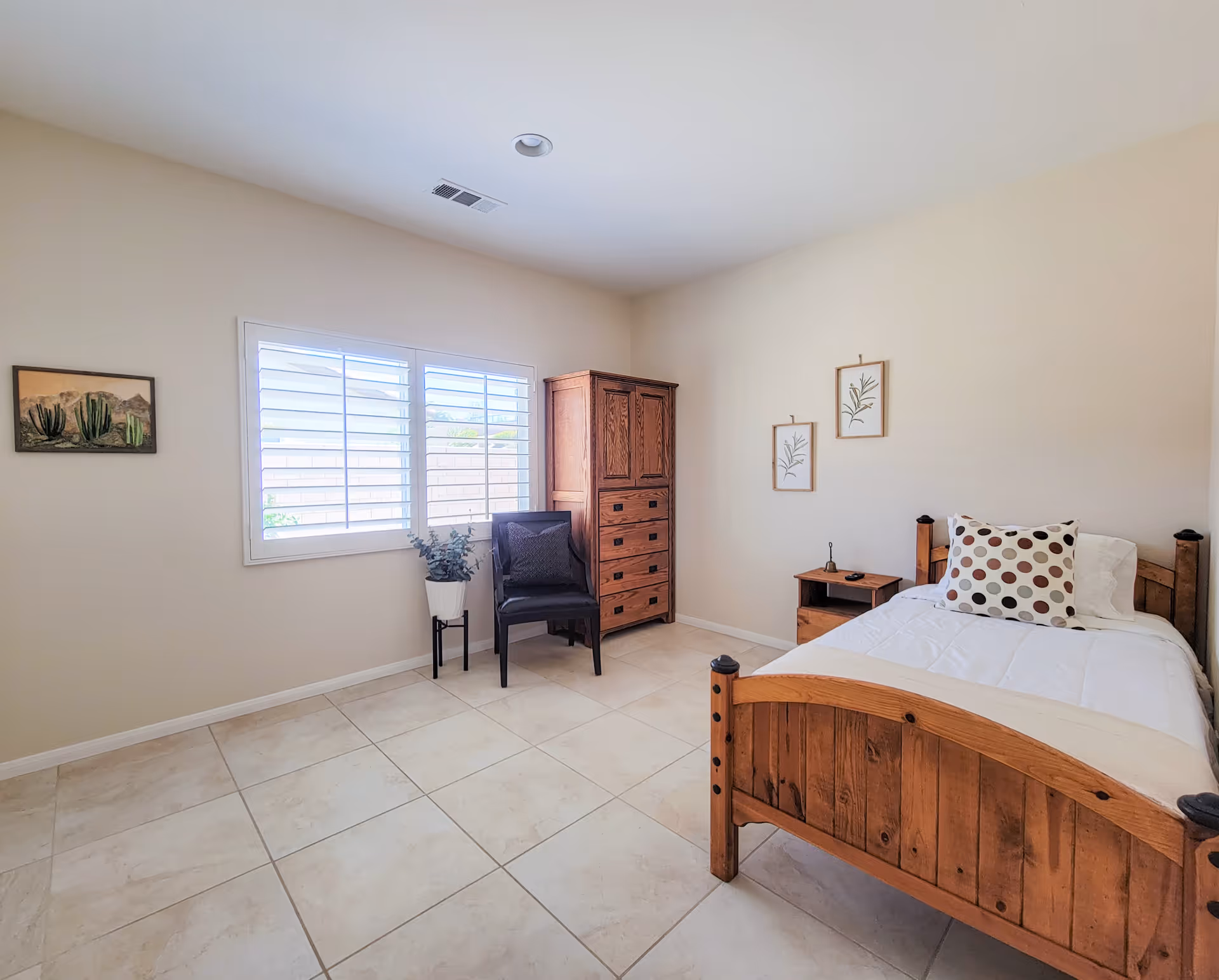 Bright, simple bedroom with a wooden single bed, nightstand, tall wooden dresser, chair, potted plant, and a window with shutters.