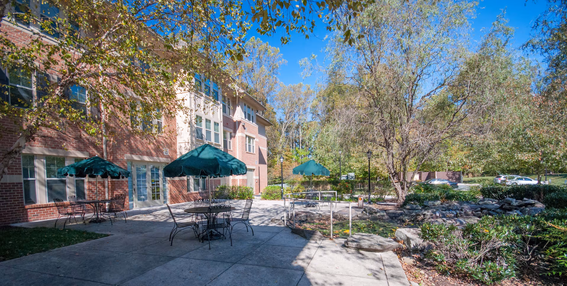 Outdoor patio area at Victory Terrace (Seniors 62+) featuring metal tables and chairs with green umbrellas, surrounded by trees and greenery, adjacent to a brick building under a clear blue sky.