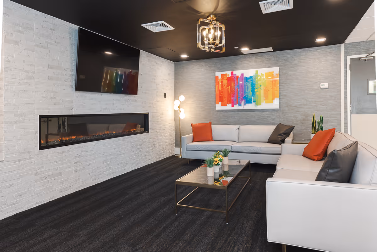 A modern living room with two light gray sofas adorned with orange and dark gray pillows, a glass coffee table with small plants, a long horizontal electric fireplace set in a white brick wall, a mounted flat-screen TV above the fireplace, a colorful abstract painting on the gray textured wall, a floor lamp with multiple round bulbs, and a black ceiling with recessed lighting and a decorative light fixture.