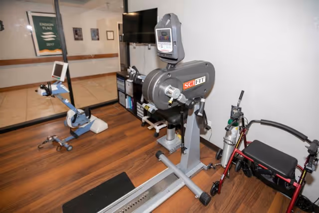 A small exercise room with two pieces of exercise equipment, including a SciFit stationary bike and another machine, on a wood floor. A red walker with a black seat is positioned near the wall. A large mirror and a TV mounted on the wall are visible in the background.