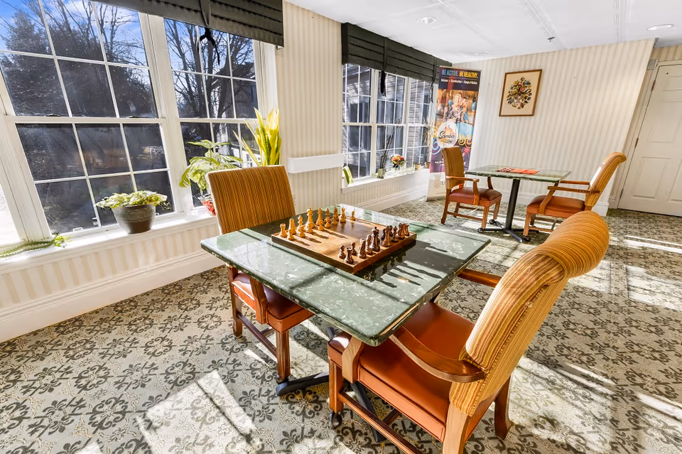A bright room with large windows letting in natural light, featuring two tables with chairs. One table has a chessboard set up for a game. The room has patterned carpet flooring, potted plants on the windowsill, and a poster on the wall promoting active living.