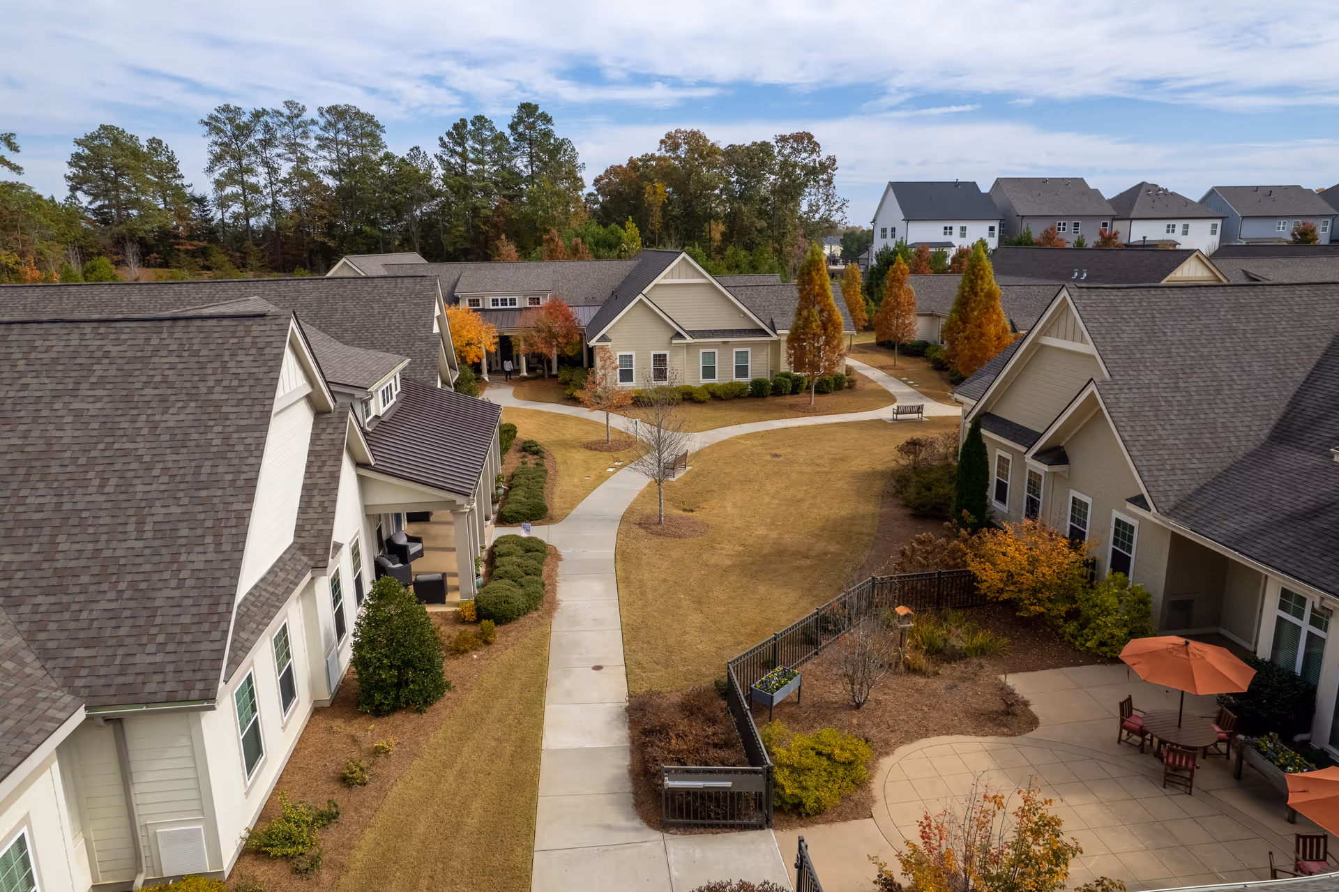 Aerial view of a senior living community courtyard with beige buildings featuring gray roofs surrounding a central lawn area with paved walkways, benches, and landscaped bushes and trees showing autumn colors. There is a small patio area with round tables, chairs, and orange umbrellas on the right side.