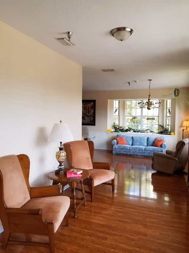 Bright communal living area with wooden floors, upholstered chairs, a blue sofa by a large window and a chandelier.
