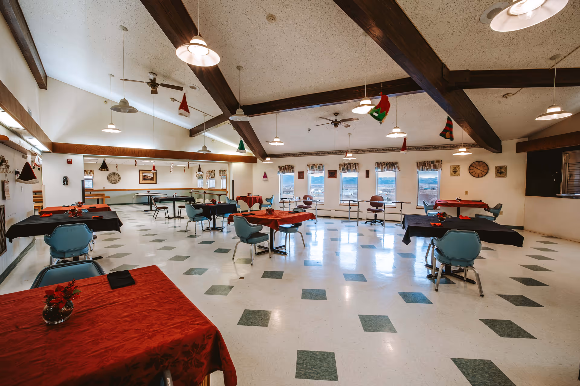 A spacious dining room with multiple tables covered in red and black tablecloths, each surrounded by blue chairs. The room has a high ceiling with exposed wooden beams and hanging pendant lights. Several windows along one wall let in natural light, and holiday decorations such as small hanging stockings and hats are visible. The floor has a white and green checkered pattern.