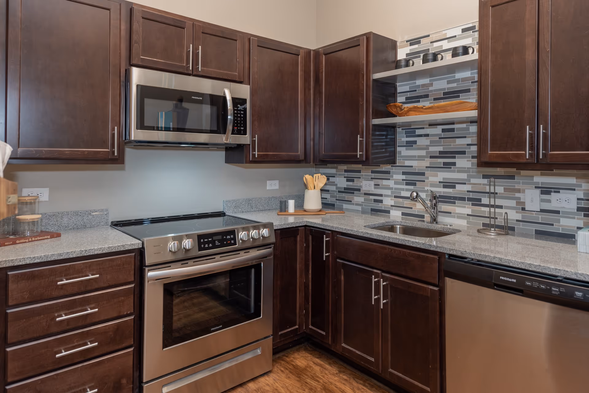 Modern kitchen with dark wood cabinets, stainless steel appliances including a microwave, oven, and dishwasher, a granite countertop, a small sink, and a tiled backsplash in shades of gray, white, and beige. There are wooden utensils in a white holder and some decorative items on open shelves.