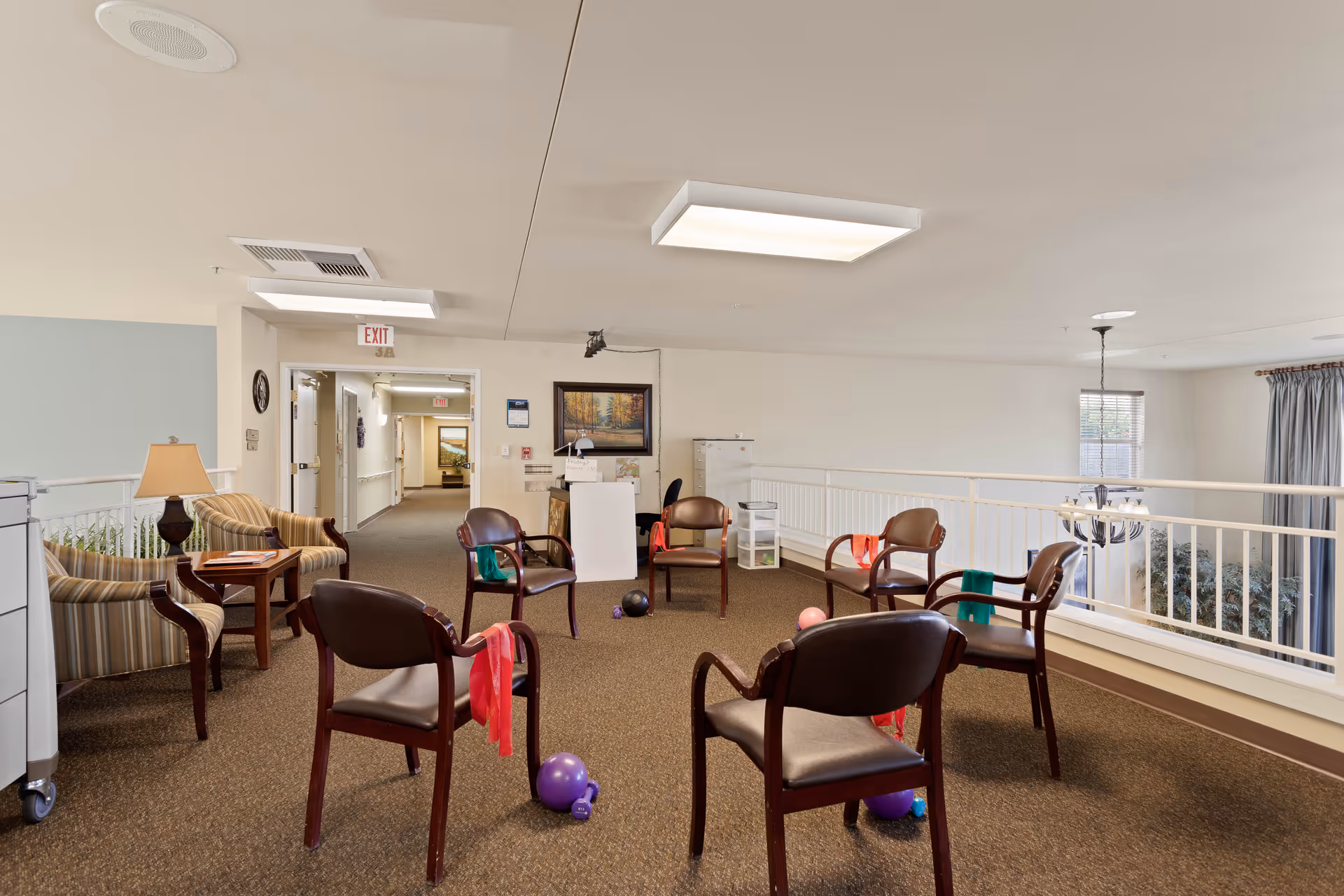 A senior living facility common area with seven chairs arranged in a circle, each with colorful resistance bands draped over them. Various exercise balls are on the carpeted floor inside the circle. The room has neutral-colored walls, a lamp on a side table, and a hallway leading to other rooms. A painting and some storage cabinets are visible in the background.
