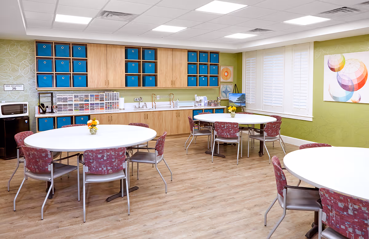 A bright and clean activity room with three round white tables surrounded by red patterned chairs. The room has light wood flooring, green walls, and a wall of cabinets with blue storage bins. There is a microwave and a small refrigerator on the left side, a sink in the middle, and colorful artwork on the walls. A window with white shutters lets in natural light.