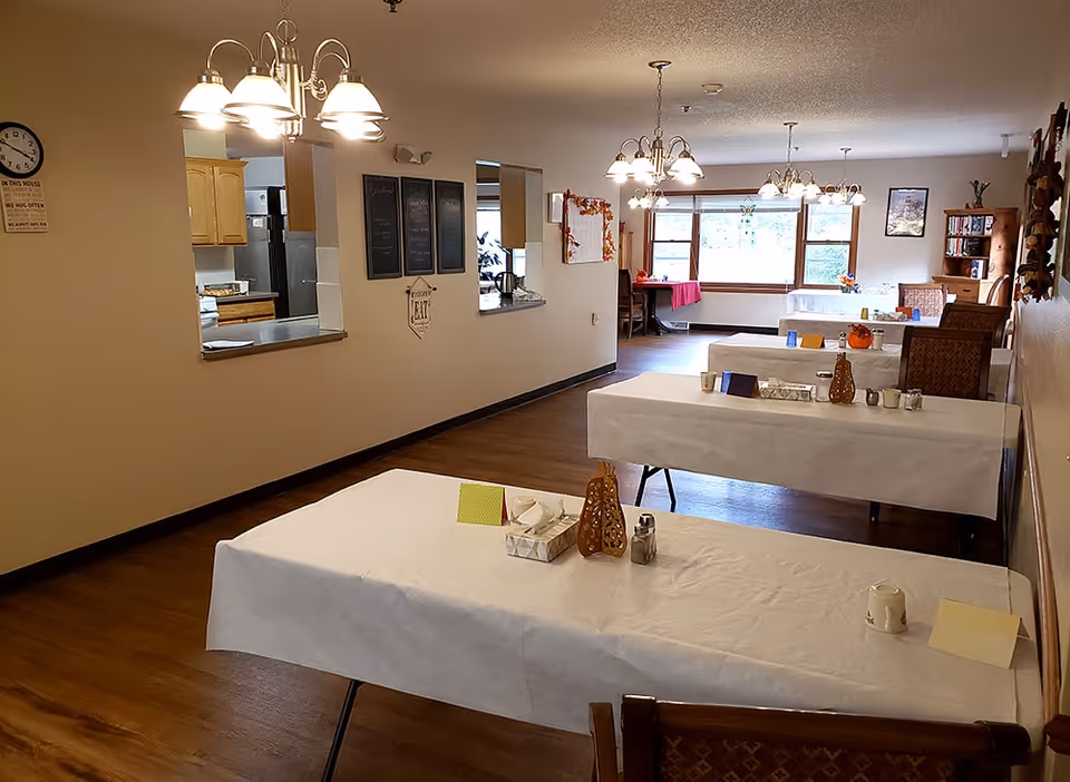 Communal dining room with several white-clothed tables, chairs, pendant lights, and a kitchen pass-through.