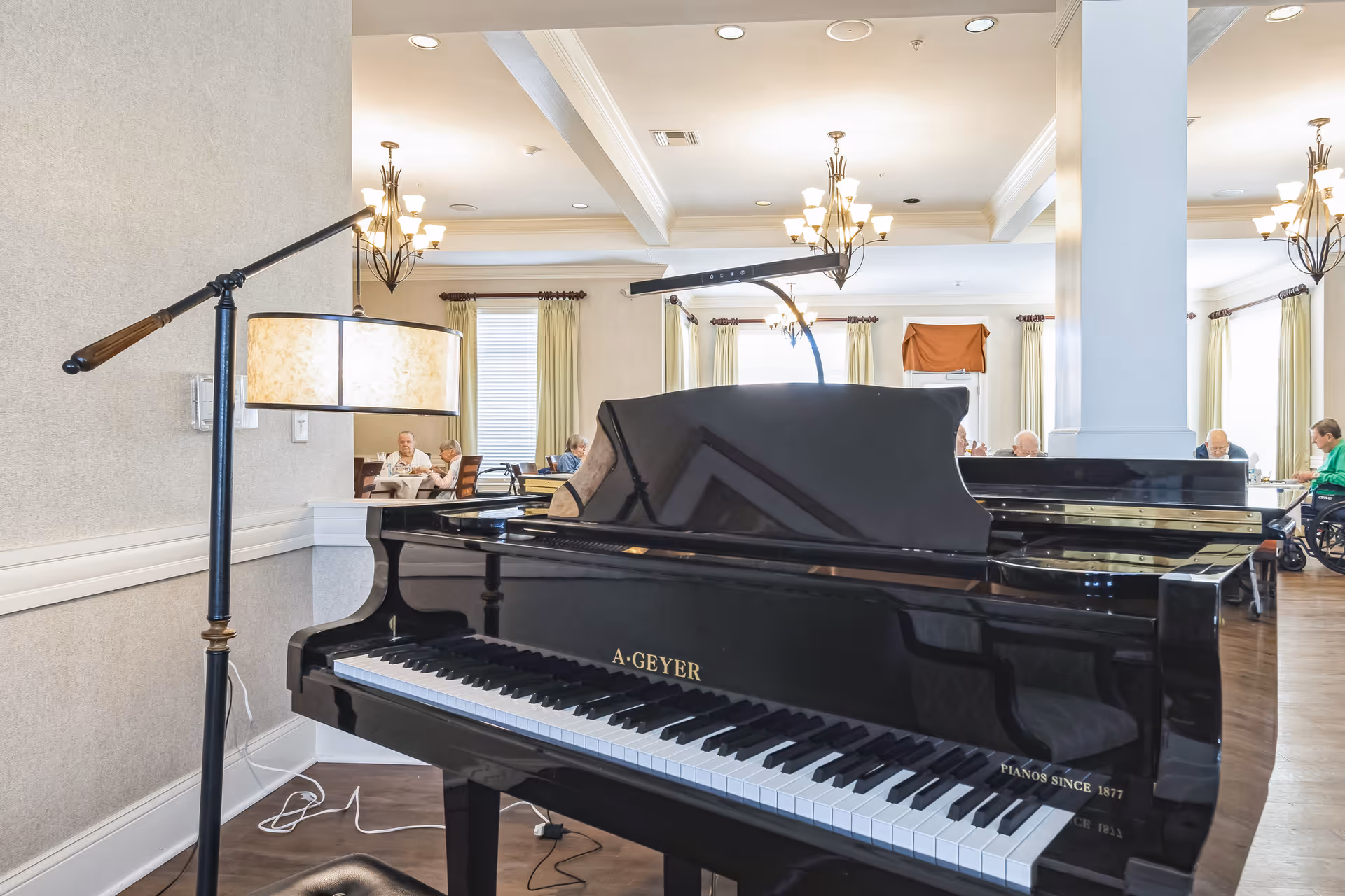 A black grand piano with the brand name A. Geyer in a well-lit room with chandeliers and large windows with curtains. Several elderly people are seated at tables in the background, some in wheelchairs, suggesting a communal or dining area in a senior living facility.