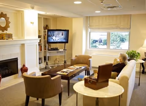 A cozy living room in a senior living facility with a woman sitting on a white sofa watching a television mounted on a stand. The room features a fireplace with decorative items on the mantel, a coffee table with magazines, two armchairs, a side table with a wooden box, a large window with a view of greenery outside, and soft lighting from wall sconces and a table lamp.