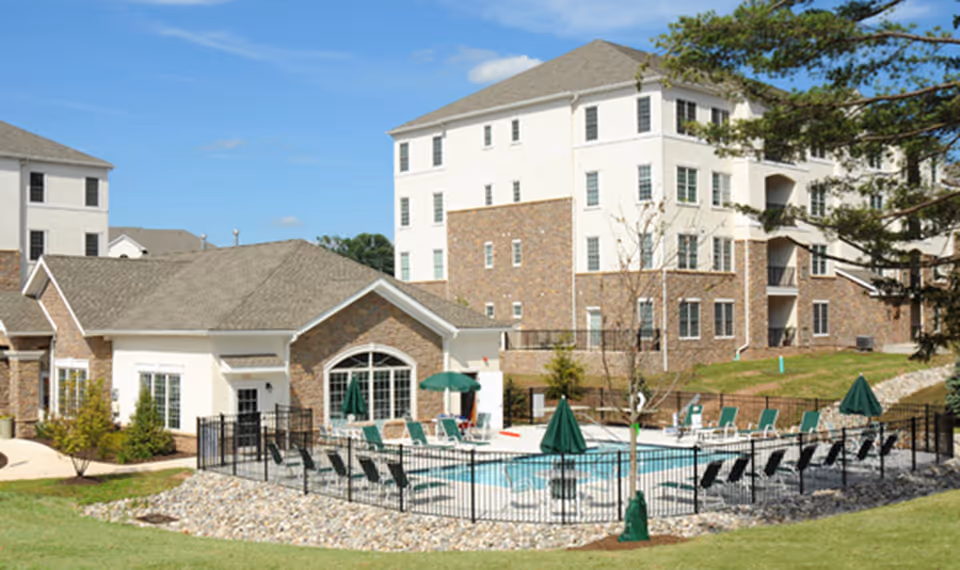 Outdoor view of a senior living facility named Dublin Terrace featuring a fenced swimming pool area with lounge chairs and umbrellas, surrounded by multi-story residential buildings and green lawn under a clear blue sky.