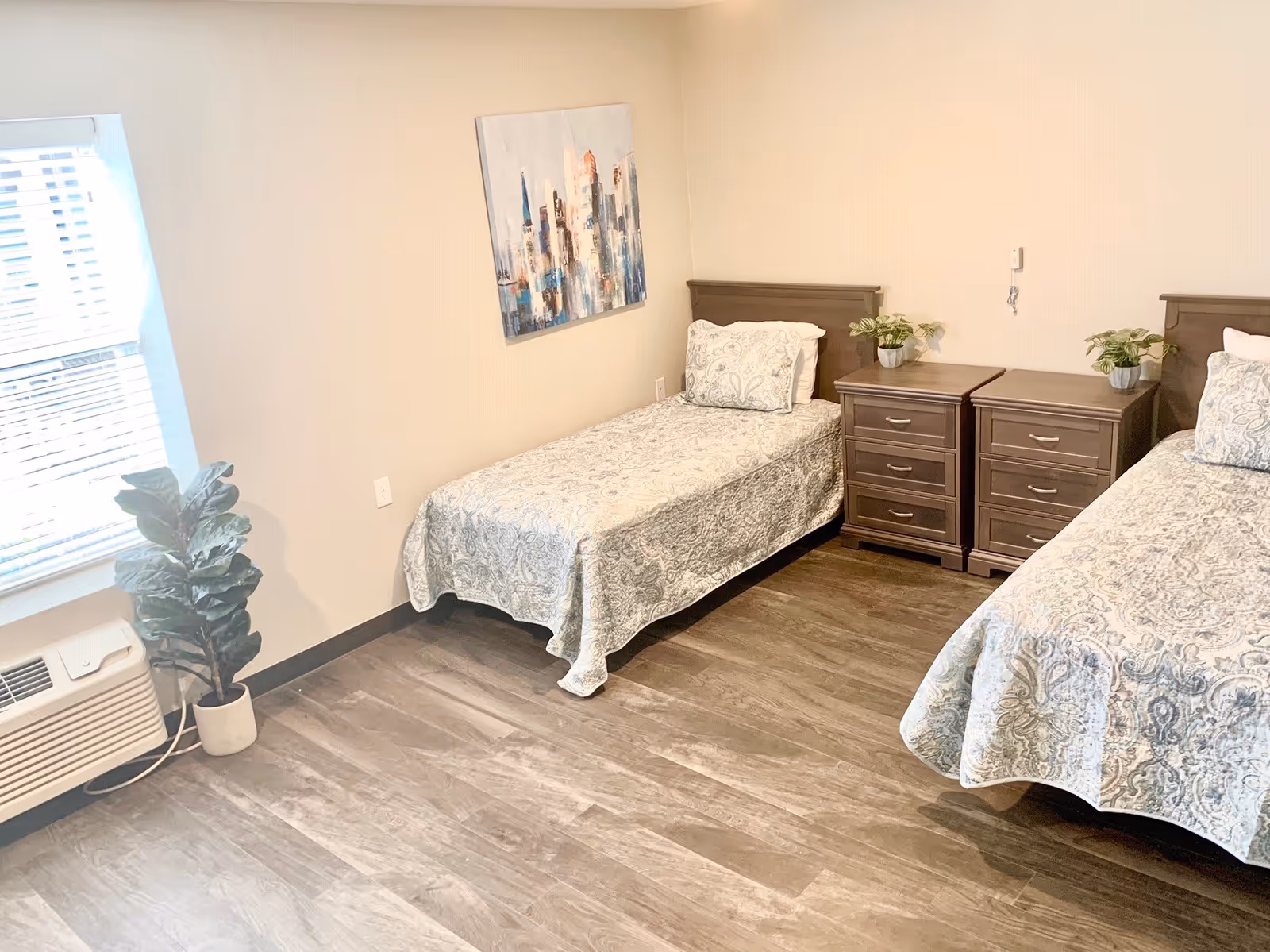 A bright bedroom with two single beds covered in matching patterned bedspreads. Between the beds are two wooden nightstands with small potted plants on top. A window with blinds and a potted plant are on the left side of the room. A painting depicting a cityscape hangs on the wall above one of the beds. The floor is wood with a light finish.