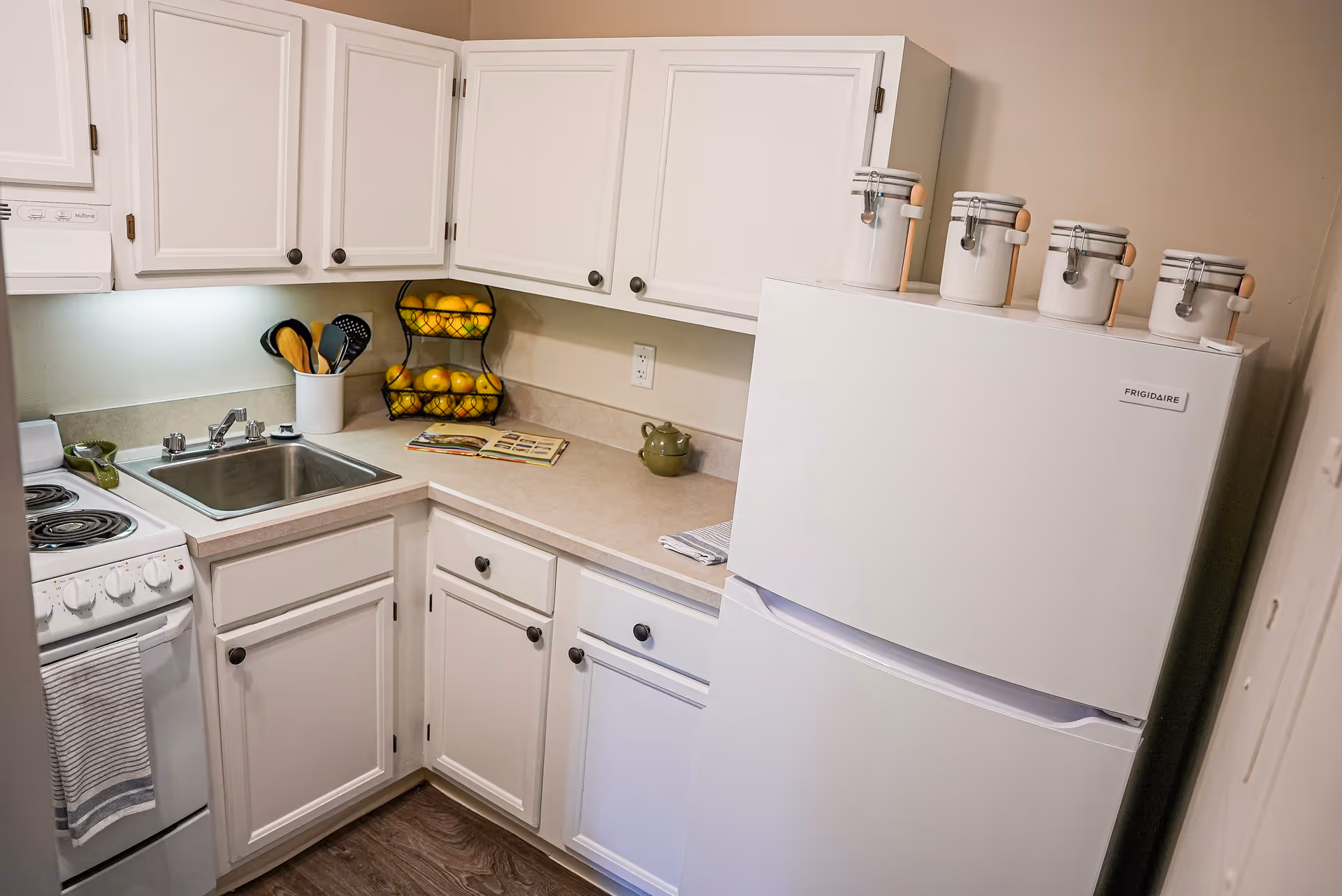 Small kitchen with white cabinets, a white refrigerator with four white canisters on top, a white stove with a striped towel hanging on the handle, a stainless steel sink, and a two-tiered fruit basket filled with lemons and apples on the countertop.