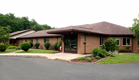 Exterior view of a single-story brick building with a brown roof, surrounded by green shrubs and trees, with a paved driveway in front.
