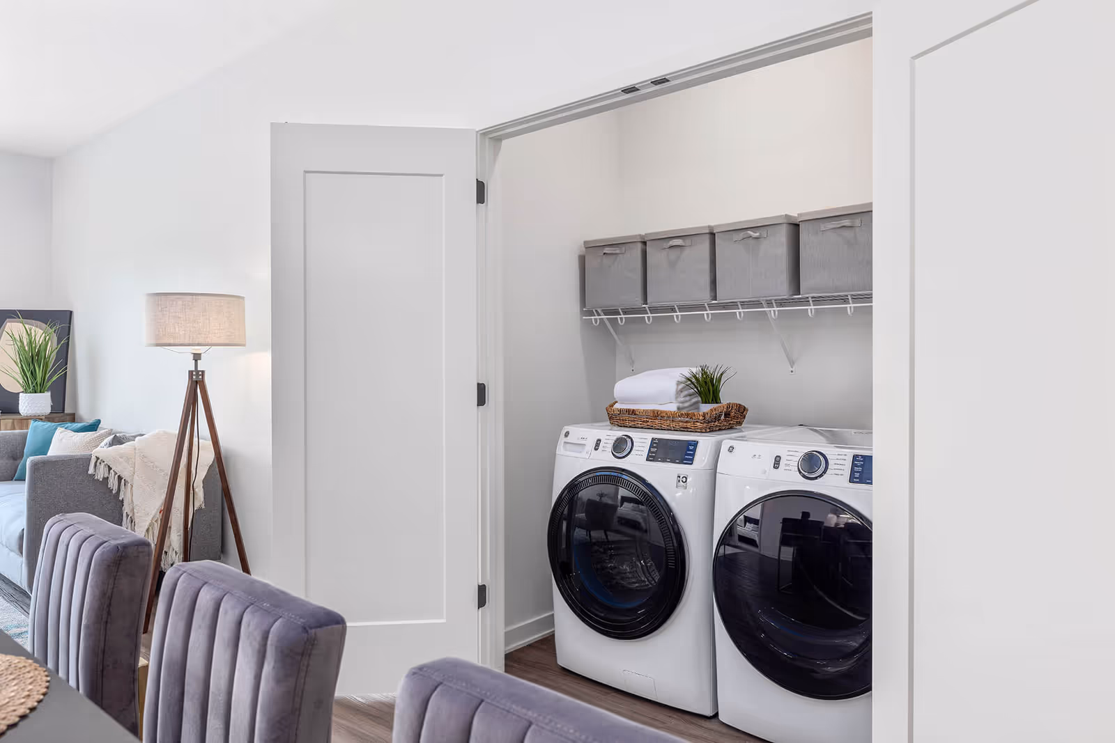 Laundry area with a white front-loading washing machine and dryer side by side inside a closet with open white double doors. Above the machines is a wire shelf holding five gray storage bins. A wicker basket with a folded white towel and a small green plant sits on top of the washing machine. In the foreground, there are purple upholstered dining chairs and part of a dining table. To the left, a gray sofa with cushions and a beige throw blanket is visible along with a wooden tripod floor lamp with a beige lampshade and a framed plant artwork on the wall.