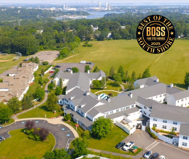 Aerial view of a senior living facility with multiple connected buildings surrounded by green lawns, trees, and parking areas. In the background, there is a large open field and a distant cityscape near a body of water. A circular driveway is visible in the foreground.