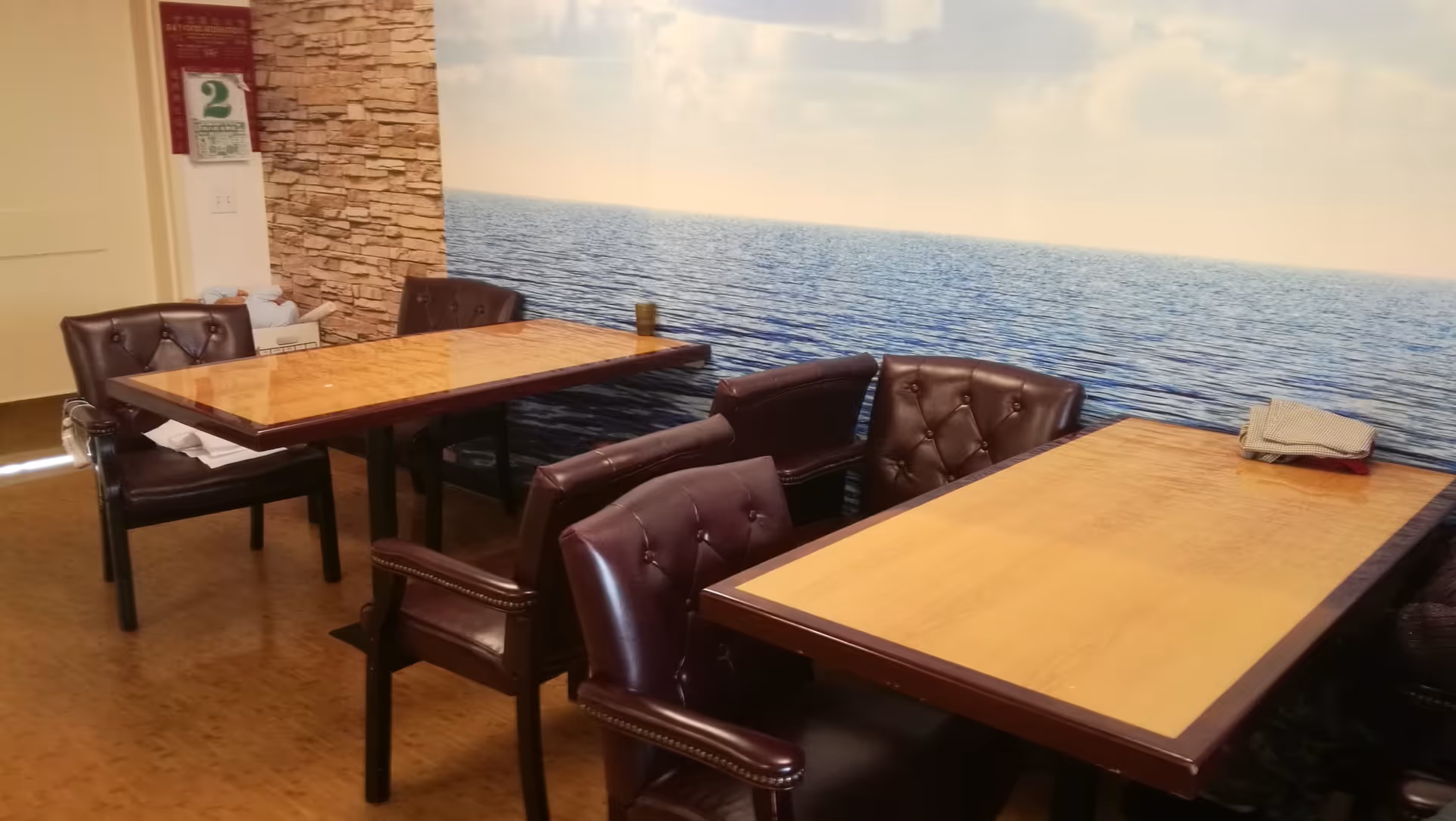 Interior room with two wooden tables and several dark brown cushioned chairs. The wall features a large mural of a calm ocean under a partly cloudy sky. There is a stone accent wall and a door in the background.